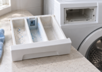 Cassetto del detersivo estratto da una lavatrice spenta, con residui visibili negli scomparti e un panno accanto
