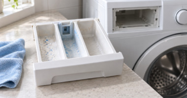 Cassetto del detersivo estratto da una lavatrice spenta, con residui visibili negli scomparti e un panno accanto